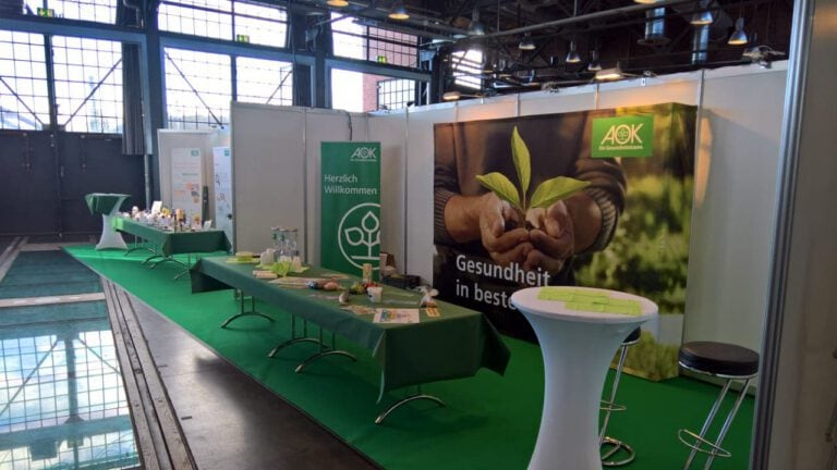Messestand mit grünen Dekorationen und AOK-Werbung bei PURA Erlebnismanufaktur.