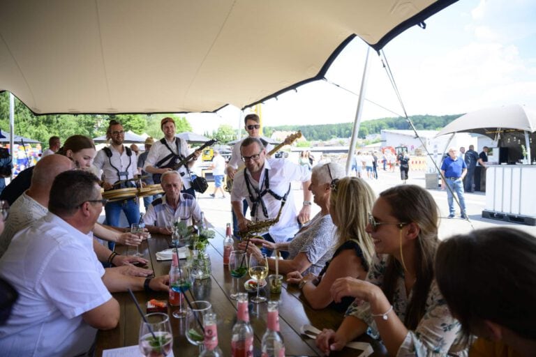 Menschen bei einem Jubiläumsfest unter einem großen Sonnensegel am Wasser.