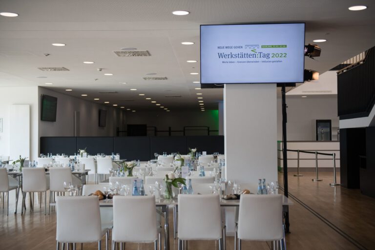 Veranstaltungsraum bei Werkstätten:Tag 2022 mit Tischen und Präsentationsbildschirm.