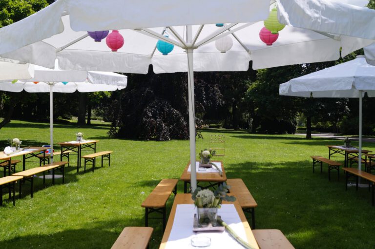 Festliche Gartenveranstaltung mit Tischen, Bänken und bunten Lampions unter Sonnenschirmen.