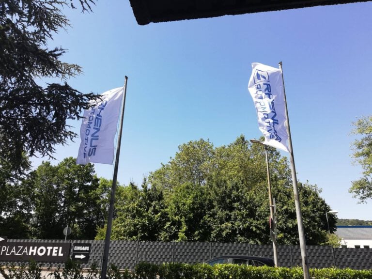PURA Erlebnismanufaktur Flaggen vor grünem Baum und blauen Himmel.